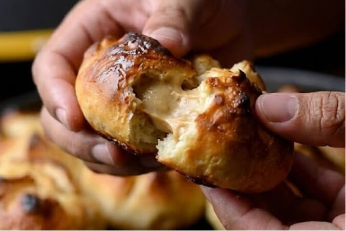 A close-up of a hand breaking open a warm Pumpkin Spice Stuffed Biscuit to reveal a creamy pumpkin filling, with more biscuits and the HALO Versa 16 pizza oven blurred in the background