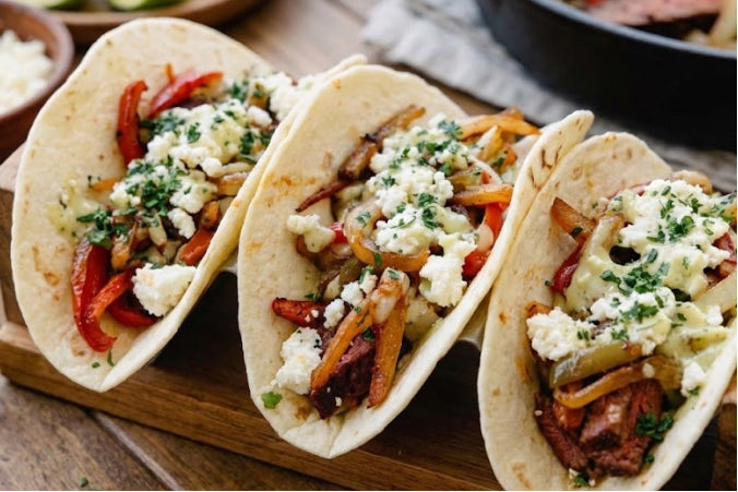 Steak tacos topped with sautéed peppers, onions, and crumbled cotija cheese on flour tortillas