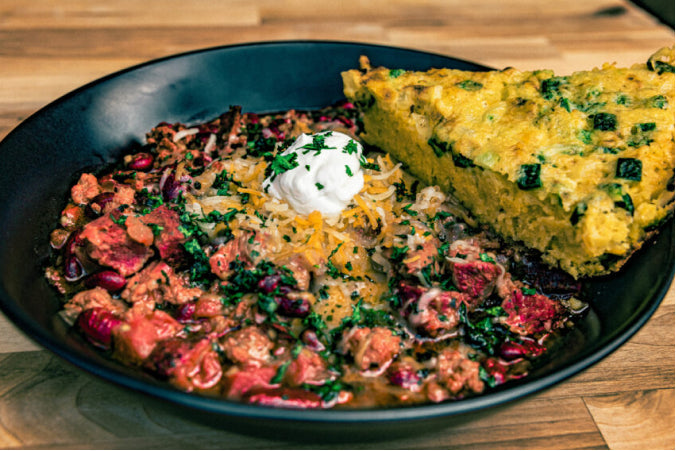 A bowl of chili featuring kidney beans and chunks of steak, topped with shredded cheese and sour cream