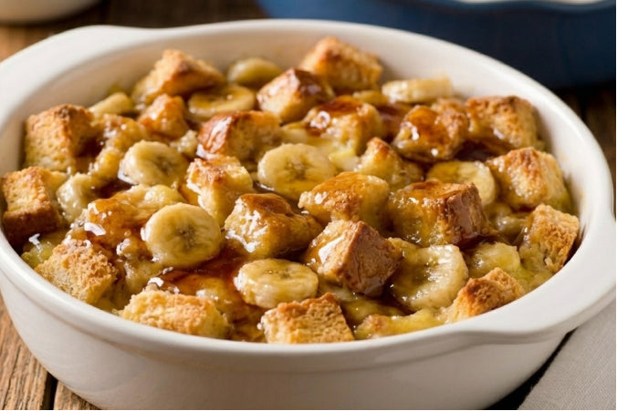 Banana Pudding in a large white bowl,  with thick banana slices and topped with a generous layer of golden-brown cubed french toast and maple syrup 