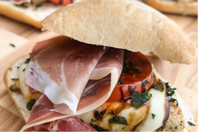 A rustic chicken caprese sandwich featuring grilled chicken, melted fresh mozzarella, a ripe tomato slice, and fresh basil, finished with a balsamic glaze drizzle and a thin slice of prosciutto.