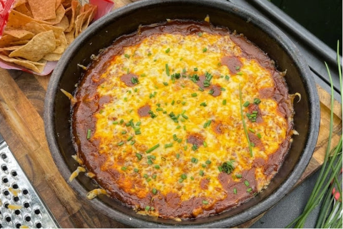 Bubbling skillet of Chili Cheese Dip with melted yellow and white cheese, topped with fresh chopped chives