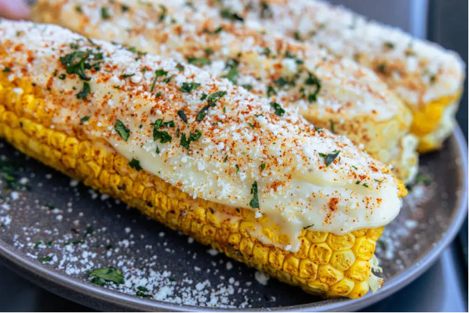 Three ears of Smoky Elote (Mexican Street Corn) prepared on a pellet grill, seasoned with chili powder and served on wooden sticks, coated in creamy mayo and crumbled Cotija cheese with fresh lime wedges on the side