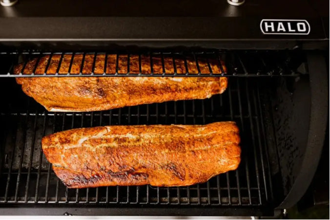 Two large salmon fillets seasoned with a sweet and spicy rub of brown sugar and bourbon smoked chili powder, smoking on the black wire grates of a HALO pellet grill