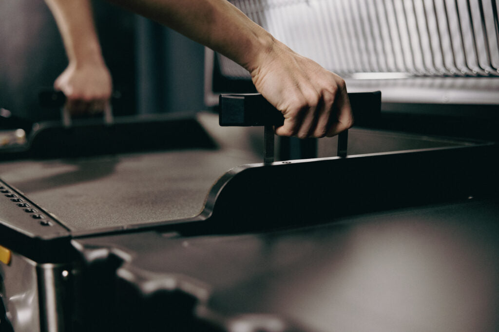 Two hands using the integrated side handles to lift and remove the heavy-duty cooking surface of a HALO Elite griddle.