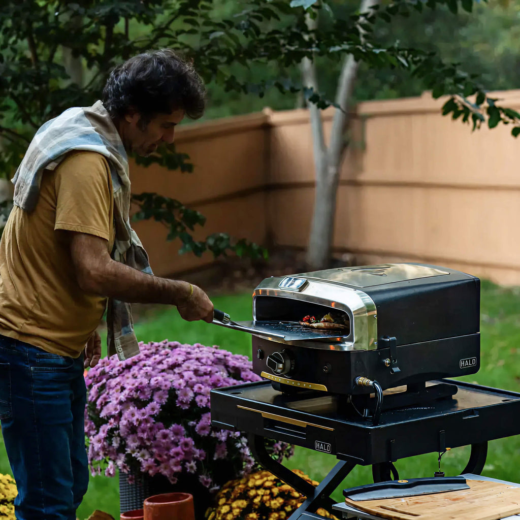 A man using a metal peel to slide a freshly baked pizza out of a HALO Versa 16 pizza oven on a sunny backyard patio