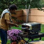 A man using a metal peel to slide a freshly baked pizza out of a HALO Versa 16 pizza oven on a sunny backyard patio