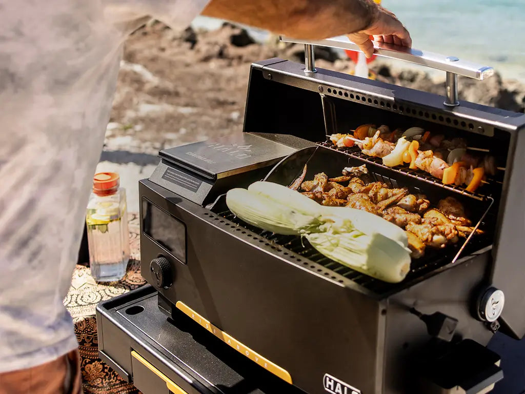 Person grilling assorted kebabs and corn on the cob on a HALO pellet grill during a sunny day at the beach