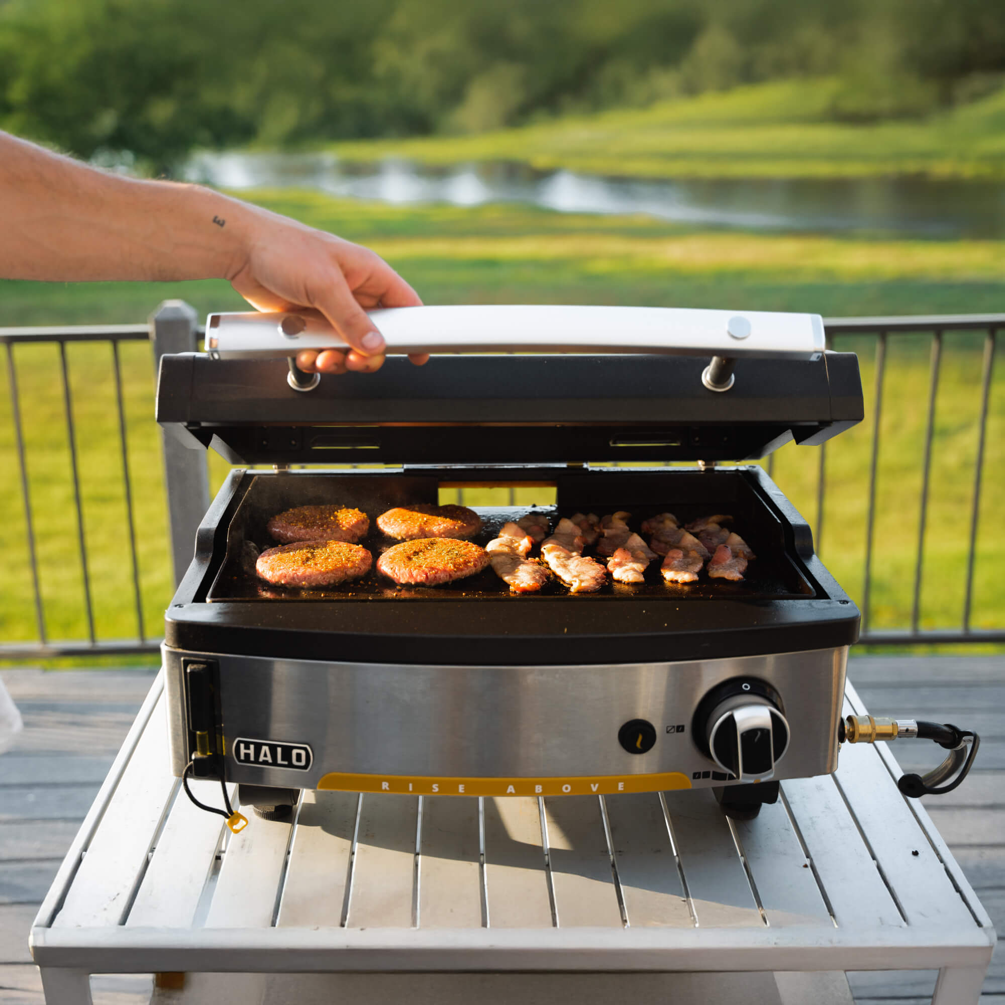 Close-up of a hand closing the lid on a HALO Elite 1B griddle with hamburgers and bacon sizzling on the cooking surface.